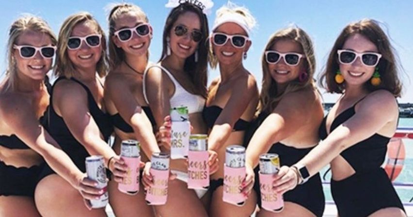 Seven women in black swimsuits and white sunglasses on a sunny boat deck, smiling and holding canned drinks in pink koozies; one person in a white tank top and visor stands at the center with blue sky and water behind them.