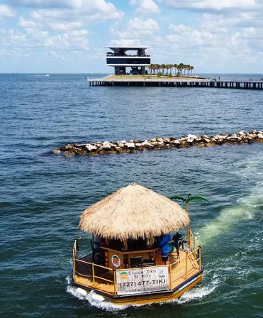 Tiki-themed thatched-roof party boat cruising across a sunny bay toward a modern waterfront pier lined with palm trees and a two-story pavilion.