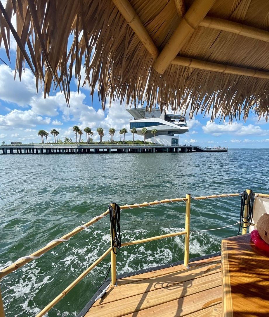 View from a thatched tiki boat deck across a sunny bay to a modern waterfront building and palm‑lined pier under blue sky with scattered clouds.