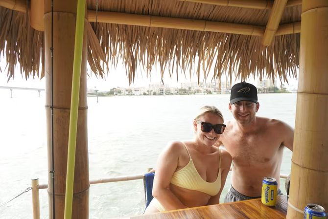 Smiling couple in swimwear at a bamboo tiki hut bar with a thatched roof, canned drinks on the wooden counter and a calm waterfront with homes across the bay — sunny vacation vibe.
