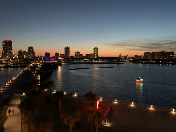 Twilight waterfront promenade with palm trees and lit pier, calm harbor reflecting a colorful illuminated boat, and downtown skyline silhouetted against a warm sunset