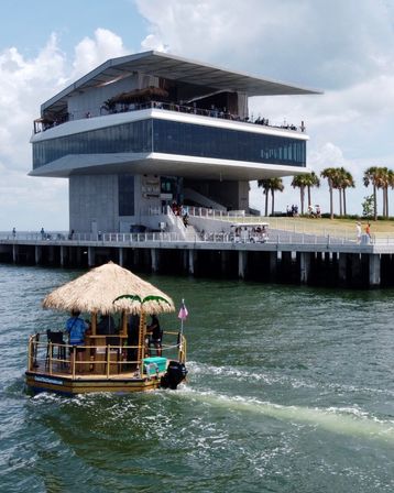 Thatched-roof tiki pontoon boat with a small American flag cruising in green water past a modern waterfront building on a palm-lined pier.