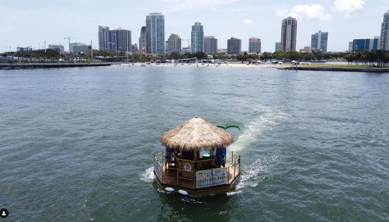 Floating tiki-bar boat cruising across harbor toward a sunny beachfront with palm trees and a modern coastal city skyline.