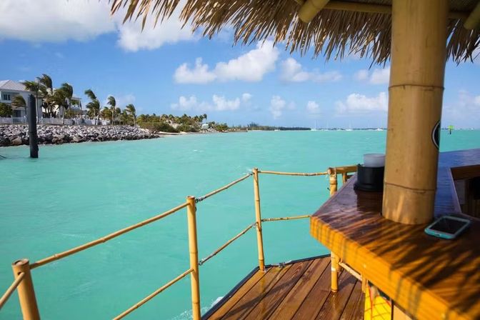 View of a turquoise tropical bay from a thatched-roof tiki bar deck with bamboo railing, palm-lined shoreline, and sailboats on the horizon