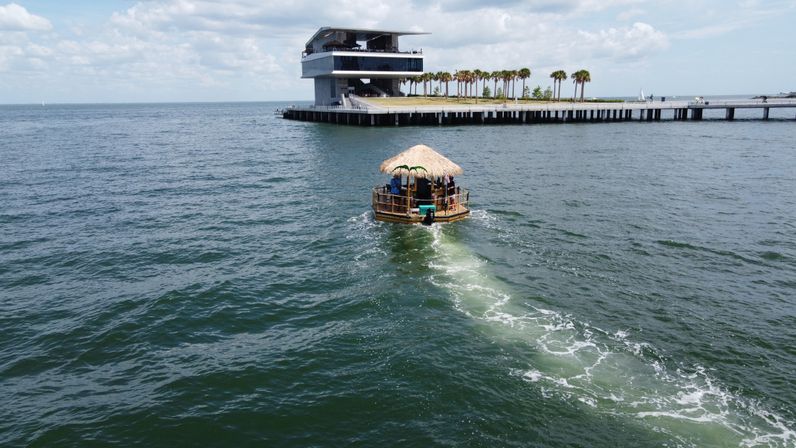 Thatched-roof tiki raft cruising across coastal waters toward a palm-lined pier and modern waterfront pavilion