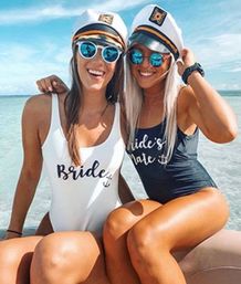 Two women in captain hats and mirrored sunglasses sit on a boat in clear shallow ocean water on a sunny day, wearing swimsuits reading 'Bride' and 'Bride's Mate'.
