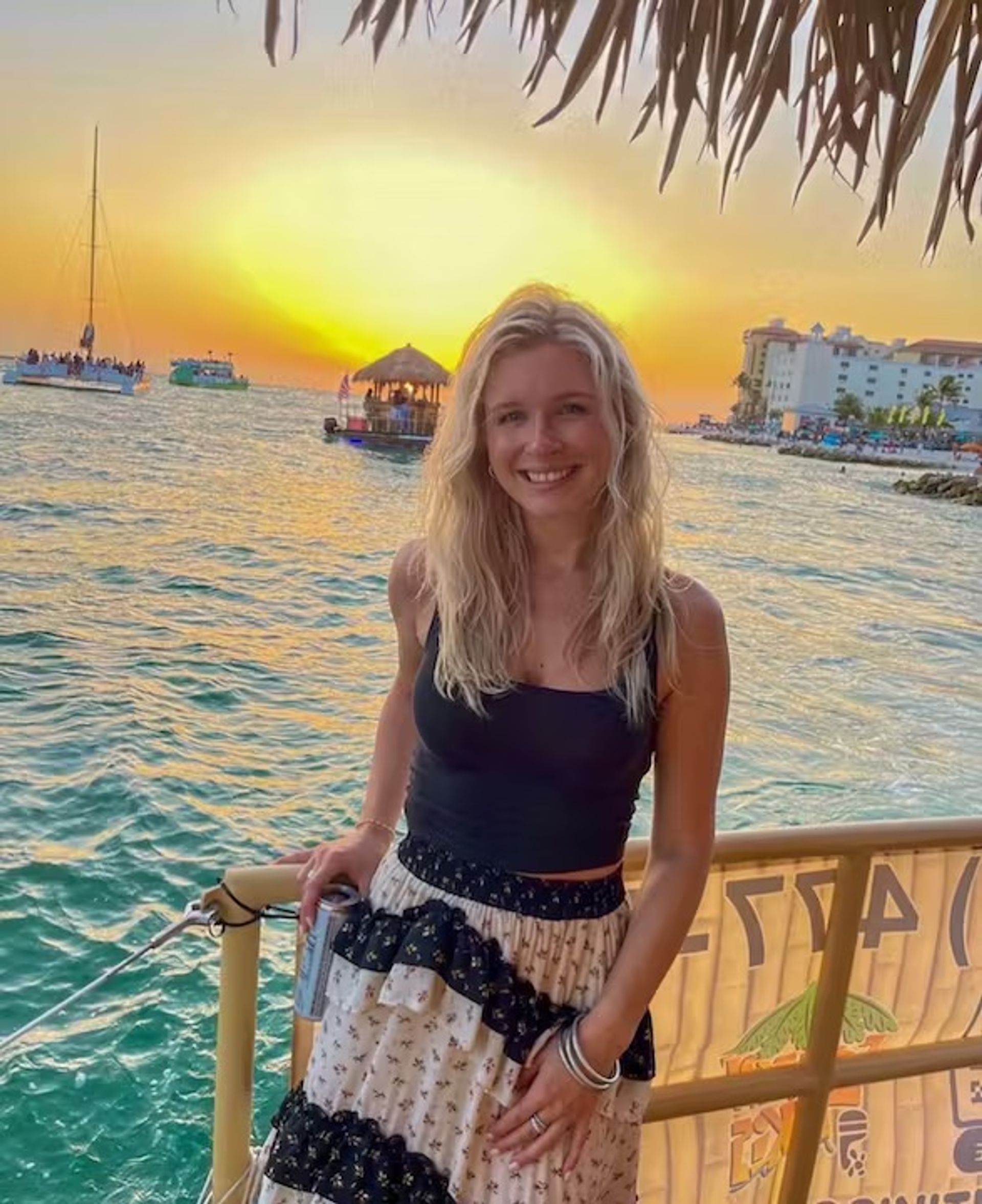 Smiling blonde woman in a black tank top and patterned skirt leaning on a railing at a tropical waterfront sunset, turquoise sea with sailboats and a thatched-roof raft in the background