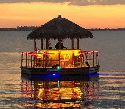 Floating tiki-bar boat with a thatched roof lit by warm neon lights, silhouetted against an orange sunset over calm bay waters with colorful reflections.
