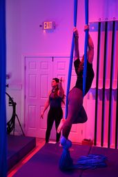 Aerial silks class in a neon-pink fitness studio: instructor guiding a student wrapped in blue silks as they climb and wrap legs above padded mats.