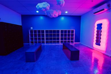 Neon-lit modern locker room in a fitness studio with open cubby shelving, two wooden benches, cloud-shaped ceiling decor and a wavy pink neon mirror, glowing in blue and magenta light.