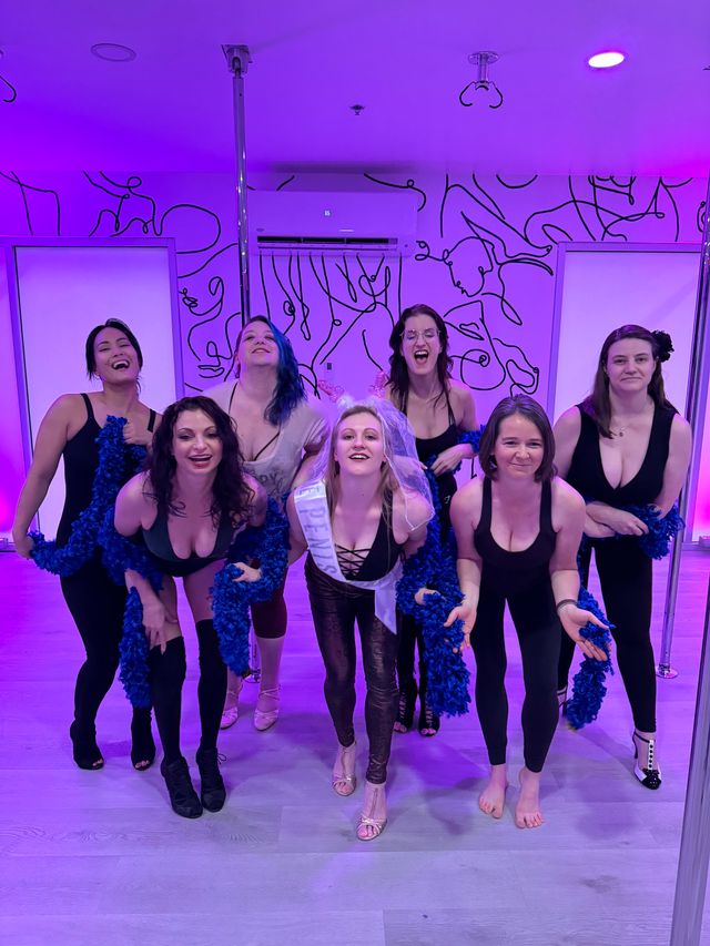Seven women posing in a purple-lit pole dance studio for a bachelorette party — bride-to-be center wearing veil and sash, all in black dancewear holding blue feather boas, smiling and playful.