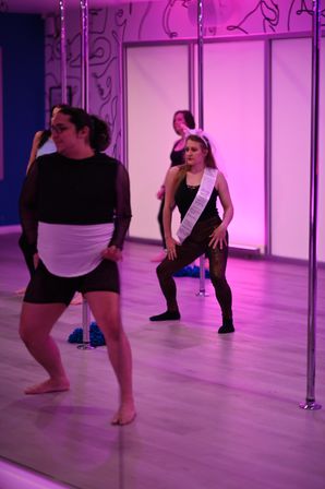 Women in a pink-lit pole fitness studio practicing dance moves; one wears a sash and veil for a bachelorette party.