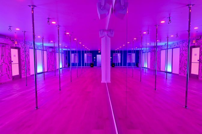 Empty neon-lit pole fitness studio with rows of chrome poles, mirrored wall reflection, hardwood floors and pink-purple ambient lighting