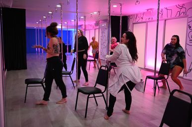 Group of women practicing pole fitness and chair choreography in a pink-lit dance studio with mirrored walls and wood-look floors