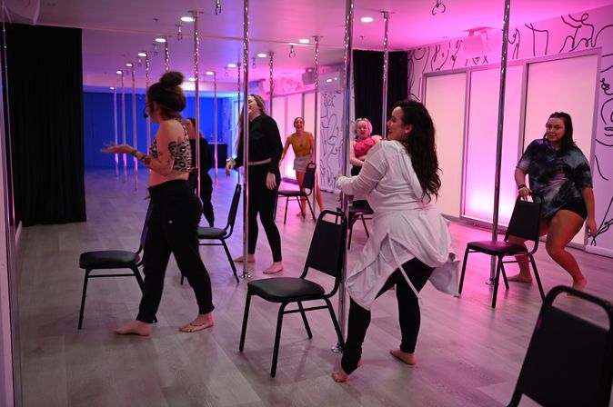 Group of women practicing pole fitness and chair choreography in a pink-lit dance studio with mirrored walls and wood-look floors