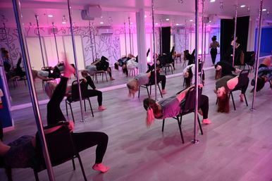 Group pole-and-chair dance class in a modern mirrored fitness studio with pink LED lighting, participants leaning back in chairs doing backbend moves on a light wood floor.