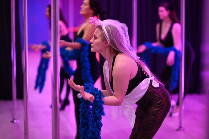Bachelorette party attendee wearing a veil and sash practices pole-dance moves in a purple-lit dance studio while holding a blue feather boa.