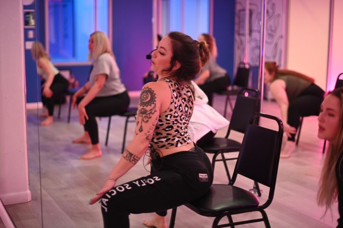 Tattooed instructor leading energetic chair-dance fitness class in a mirrored studio, students doing seated squats under pink-and-blue mood lighting.