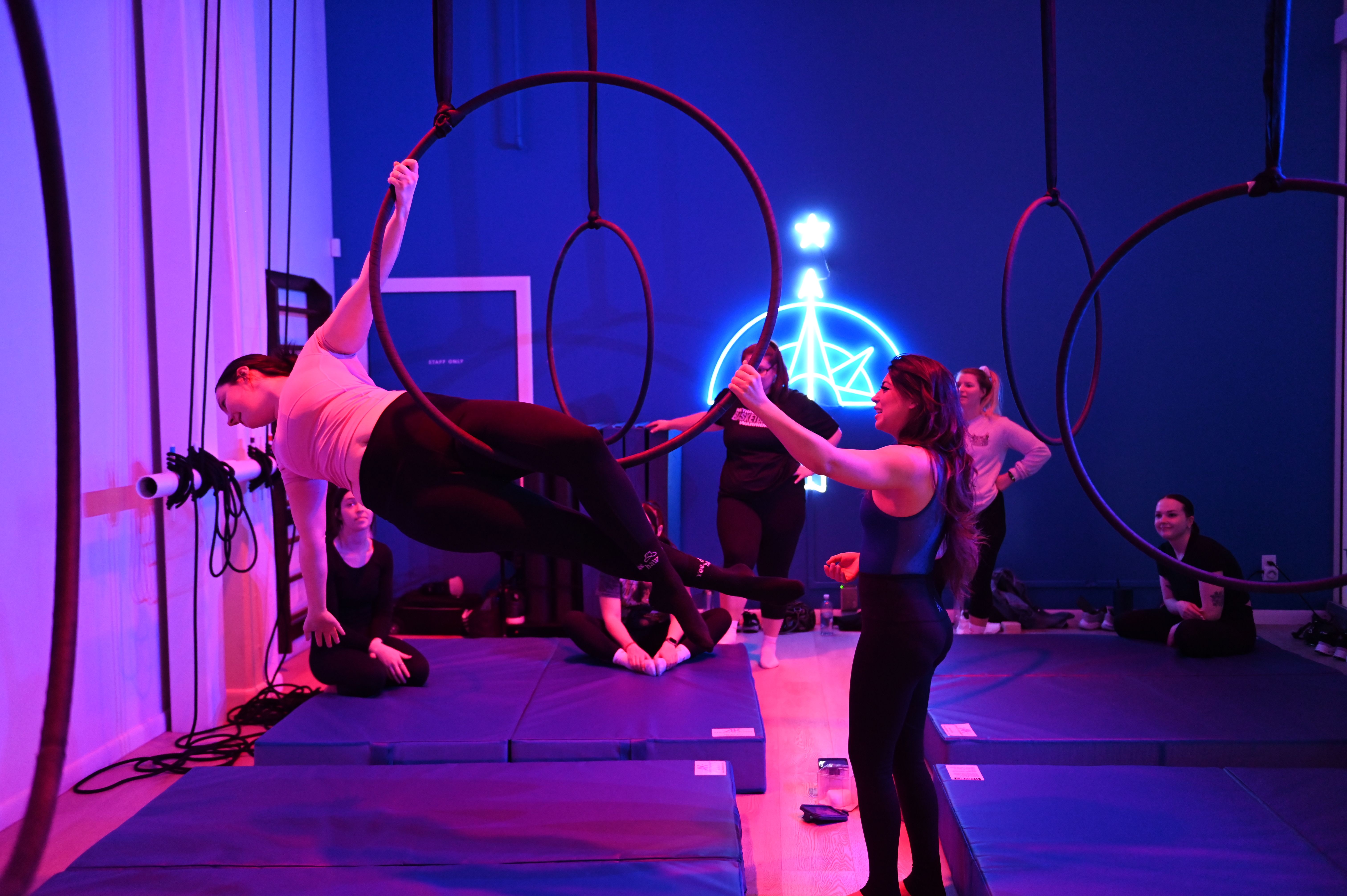 Neon-lit aerial hoop (lyra) class in an indoor studio with purple-blue lighting; instructor assists a student holding a suspended pose on a hoop while classmates sit on mats and watch.