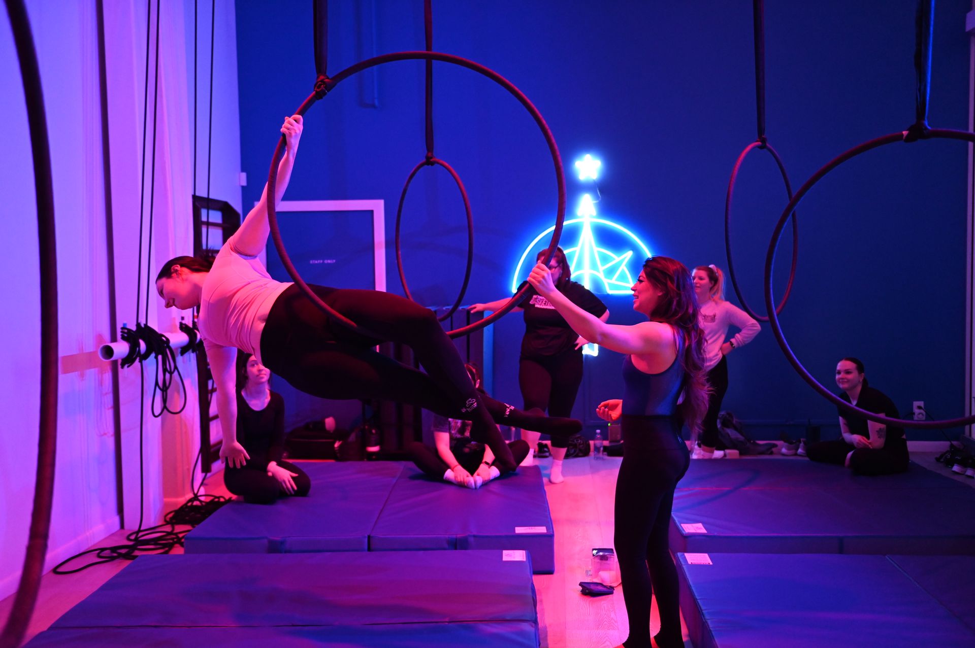 Neon-lit aerial hoop (lyra) class in an indoor studio with purple-blue lighting; instructor assists a student holding a suspended pose on a hoop while classmates sit on mats and watch.