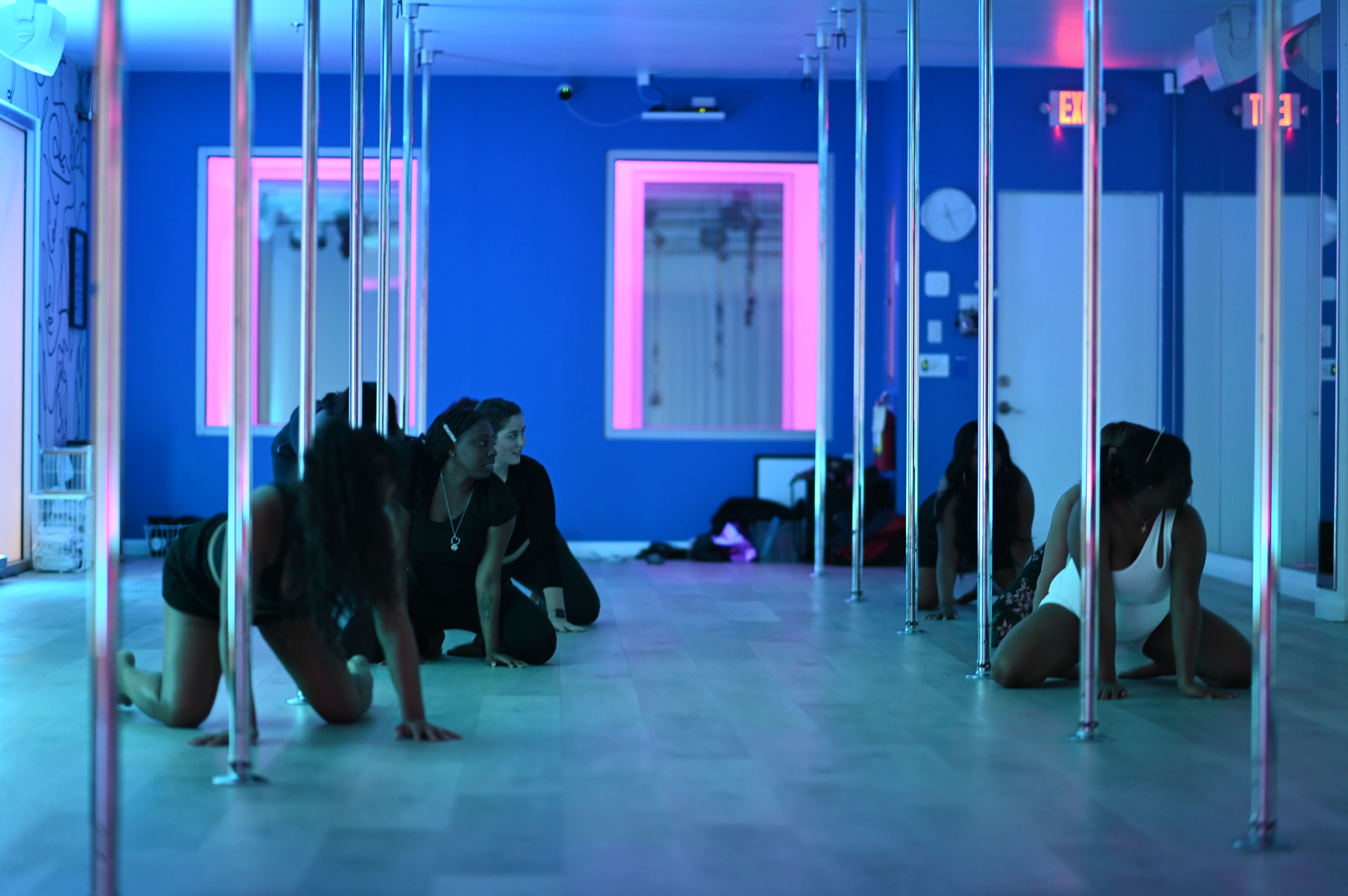 Neon-lit pole fitness class in a blue and pink indoor dance studio, participants stretching on the floor between chrome poles.