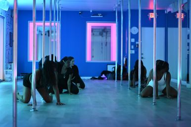 Neon-lit pole fitness class in a blue and pink indoor dance studio, participants stretching on the floor between chrome poles.
