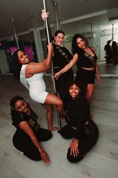 Five friends smiling and posing around a chrome pole in a modern pole-dance studio, bachelorette party vibe and group fitness fun