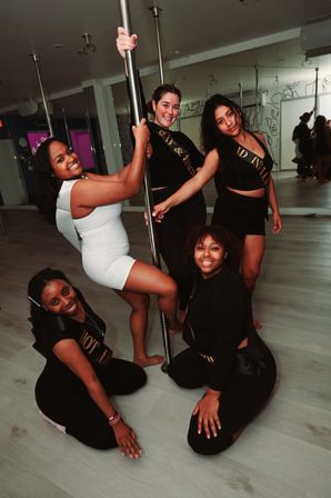Five friends smiling and posing around a chrome pole in a modern pole-dance studio, bachelorette party vibe and group fitness fun