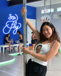 Smiling woman in workout clothes wearing a bride headband holds a chrome pole inside a bright pole-dance studio with a blue accent wall and other women practicing in the background.