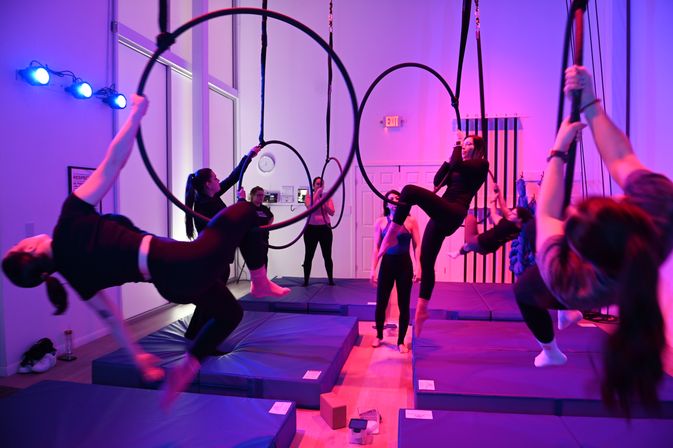 Group aerial hoop class in a purple-lit indoor fitness studio, participants practicing suspended hoop and rope poses above padded mats.