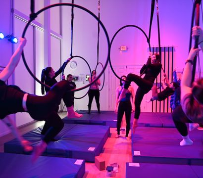 Group aerial hoop class in a purple-lit indoor fitness studio, participants practicing suspended hoop and rope poses above padded mats.