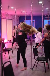 Group of women in a pink-and-blue lit pole fitness studio practicing a barefoot dance class, flipping hair beside chrome poles and chairs on a wood-look floor.