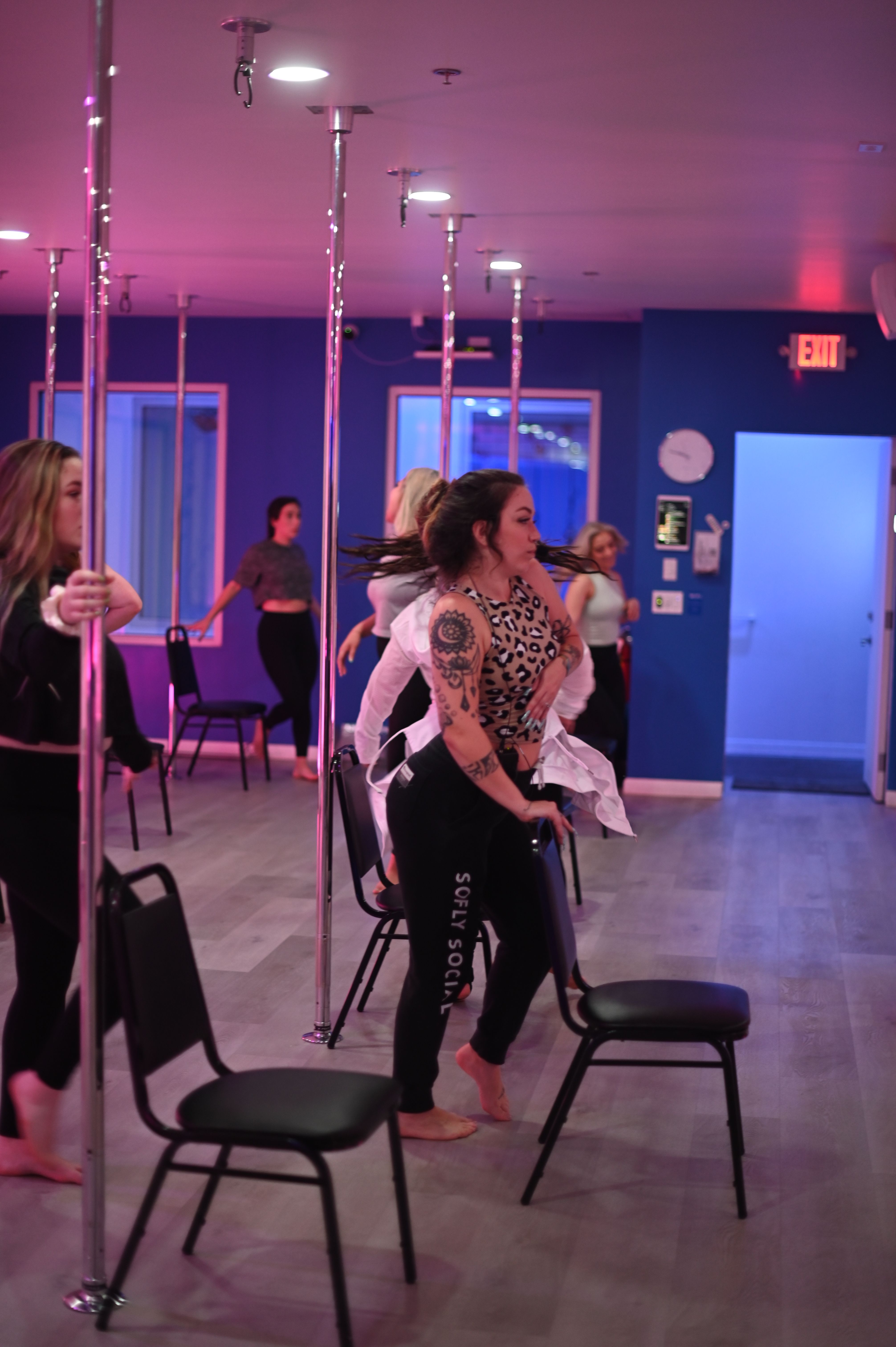 Group pole-fitness dance class in a purple-pink lit studio—women practicing chair choreography around chrome poles, tattooed dancer in foreground.