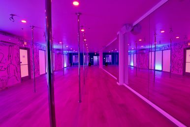 Vibrant pink-purple pole fitness studio interior with rows of chrome poles on a wood floor, long mirrored wall, neon-accented mural art and glowing exit doors.