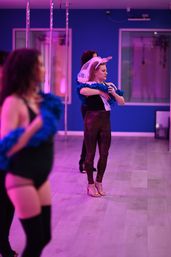 Women in a pink‑lit pole dance studio practicing with blue feather boas; one wearing a bridal veil and heels during a bachelorette dance class.
