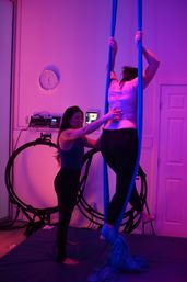 Instructor assisting a student on blue aerial silks in a purple-lit indoor aerial yoga/fitness studio with padded floor and training rings.