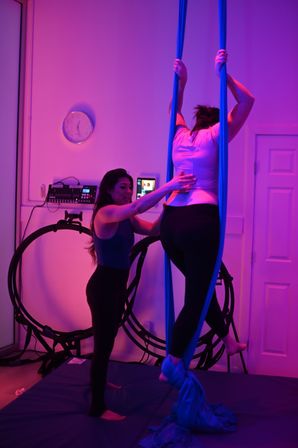Instructor assisting a student on blue aerial silks in a purple-lit indoor aerial yoga/fitness studio with padded floor and training rings.