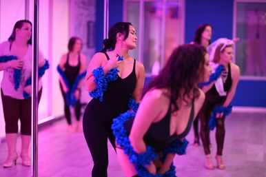 Group of women in a pink-lit dance studio practicing a playful burlesque-style routine with blue feather boas by mirrored walls and poles