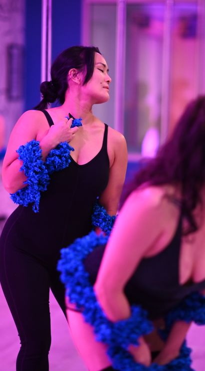 Group of women in a pink-lit dance studio practicing a playful burlesque-style routine with blue feather boas by mirrored walls and poles