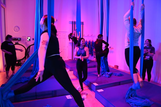 Group aerial yoga class in a neon-lit studio using blue aerial silks over padded mats, participants climbing, stretching, and spotting each other under purple-pink mood lighting