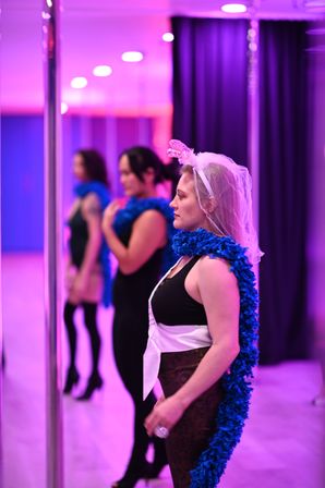 Participant wearing a veil, sash and blue feather boa practicing pole dance in a purple-lit studio, mirrored wall reflecting two blurred classmates in the background.