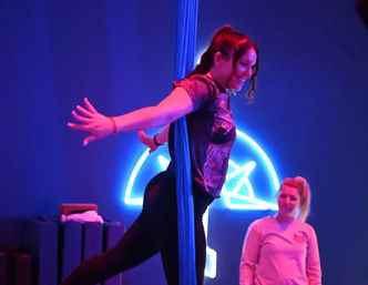 Woman practicing aerial silks pose on blue fabric in a neon-lit fitness studio while a coach watches, bathed in purple and blue lighting