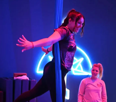 Woman practicing aerial silks pose on blue fabric in a neon-lit fitness studio while a coach watches, bathed in purple and blue lighting