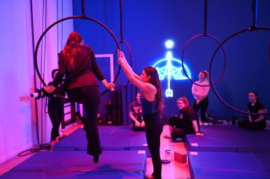 Neon-lit aerial hoop class in a fitness studio — instructor steadies a woman stepping into a suspended metal hoop while classmates watch from padded mats.