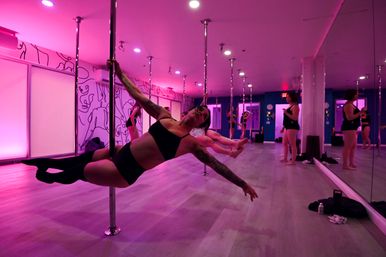Group pole fitness class in a pink-lit studio, participants practicing strength holds on chrome poles with mirrored walls and light wood floors.