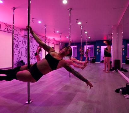 Group pole fitness class in a pink-lit studio, participants practicing strength holds on chrome poles with mirrored walls and light wood floors.