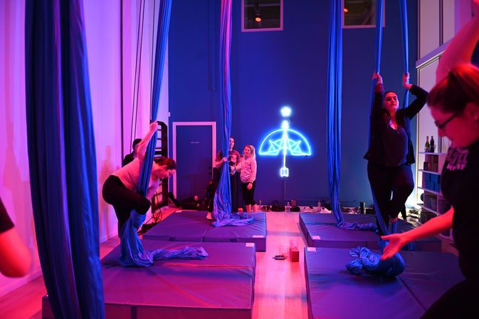 Participants practicing aerial silks in a neon-lit fitness studio — blue fabric hammocks over padded mats under purple-blue lighting with a glowing umbrella wall sign.