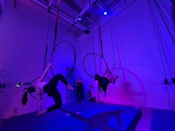 Two aerialists swinging on suspended lyra hoops in an indoor aerial hoop studio bathed in purple-blue LED lights, with padded mats below and a person filming in the background.