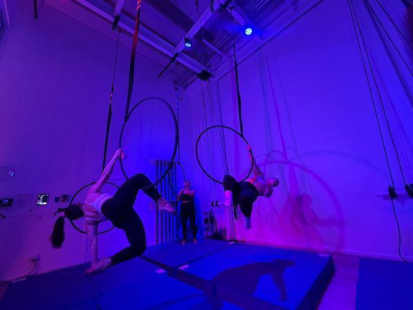 Two aerialists swinging on suspended lyra hoops in an indoor aerial hoop studio bathed in purple-blue LED lights, with padded mats below and a person filming in the background.