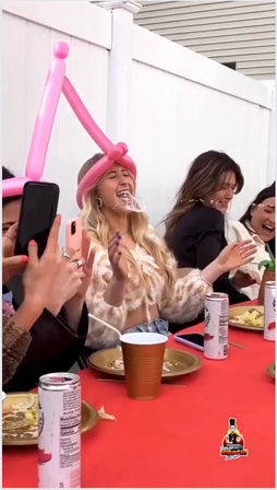 Backyard party scene with friends laughing at a red-covered table; woman wearing a pink balloon crown blows a drink-splashed bubble while others film on their phones, plates and canned drinks nearby.